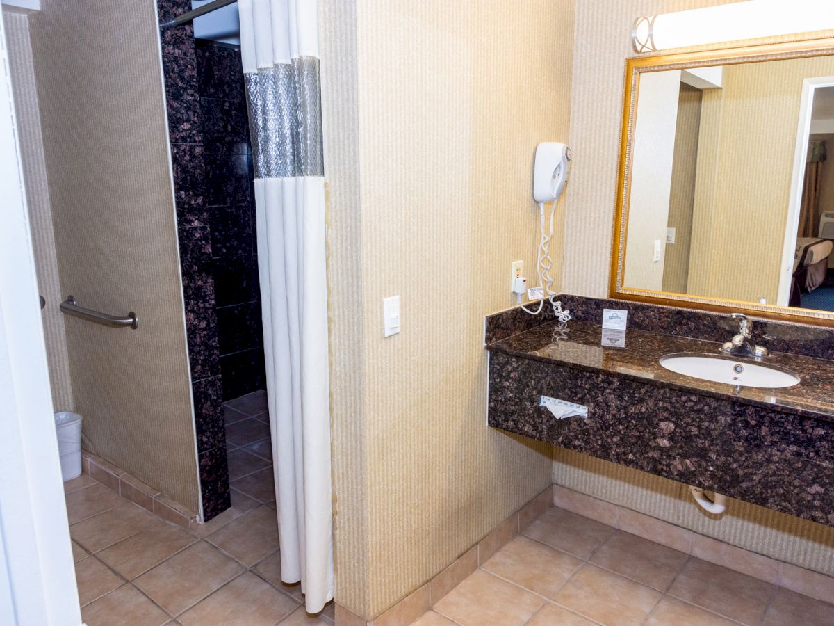 This image shows a hotel bathroom with a sink, a granite countertop, a mirror, a hairdryer, a shower area with a curtain, and a grab bar.