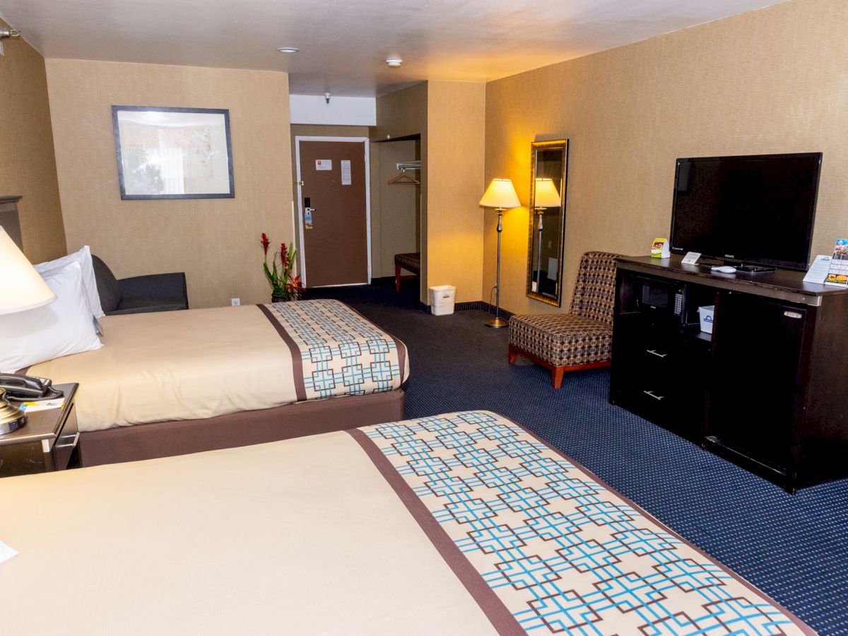 The image shows a hotel room with two beds, a TV on a dresser, a chair, and a lamp on a side table near a wall-mounted mirror.