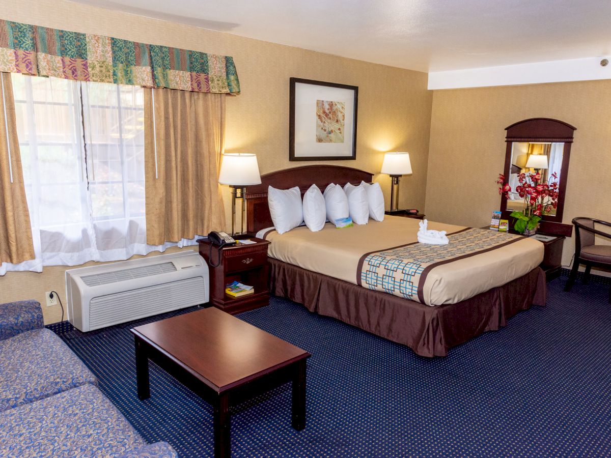 A cozy hotel room with a bed, lamps, a couch, a table, and an air conditioner by the window. Artwork and flowers add decor.