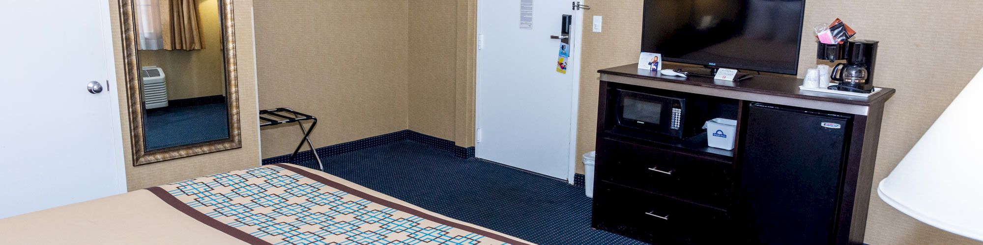 A hotel room with a bed, TV, mini-fridge, microwave, mirror, hangers, and a lamp on a table, featuring beige and blue decor, and carpeted flooring.