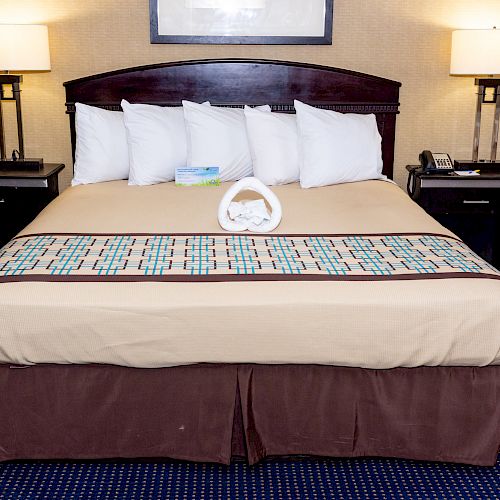 A neatly made hotel bed with a decorative runner, flanked by two bedside tables with lamps, and a towel arrangement at the center of the bed.