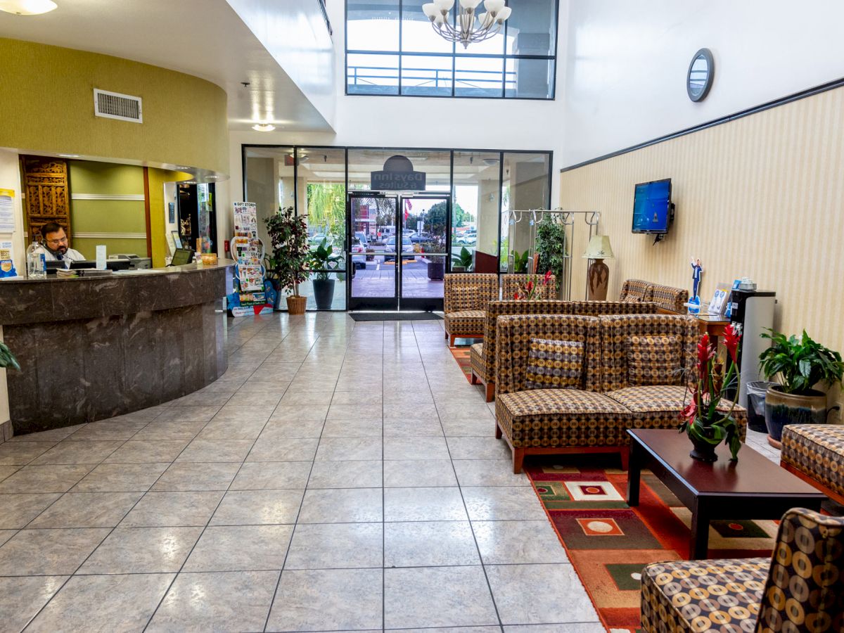 A lobby area with check-in desk, seating, plants, and a TV on the wall. The entrance and exterior view are visible at the far end of the room.