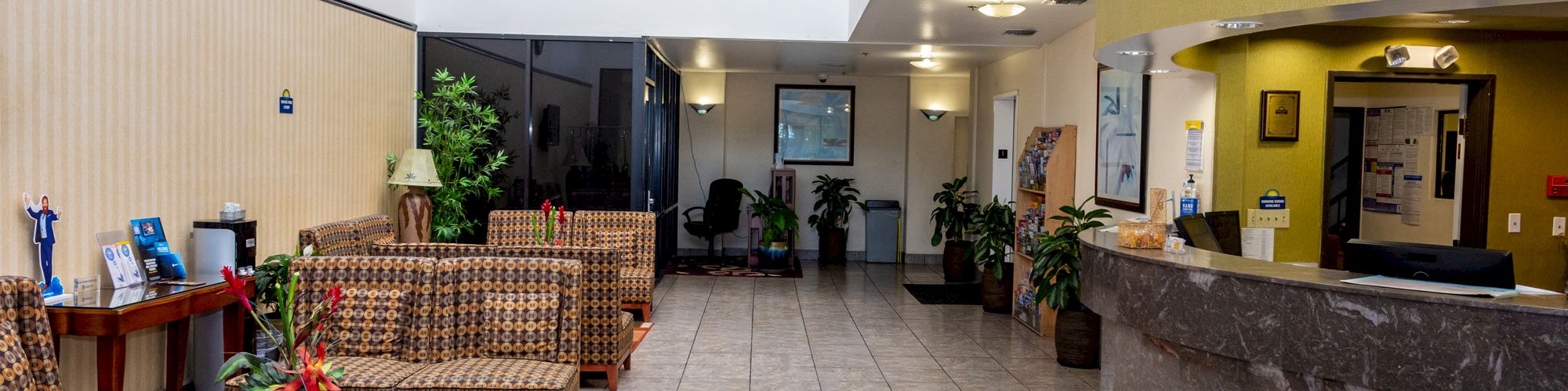 The image shows a lobby with patterned chairs, a tiled floor, a reception desk, plants, and decorations.