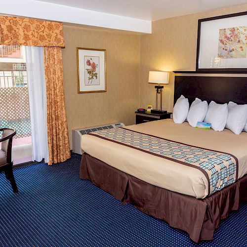 This image shows a hotel room with a large bed, five pillows, two bedside lamps, a small table with chairs, two framed artworks, and a window with curtains.