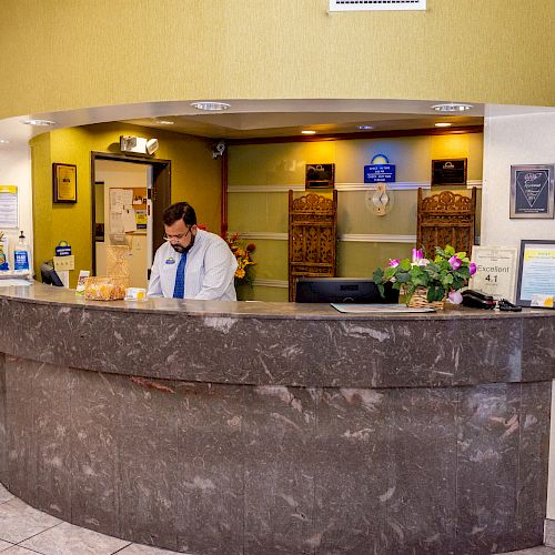 A receptionist behind a brown marble front desk in a hotel lobby with flowers, a computer, and a store selling gifts on the side.