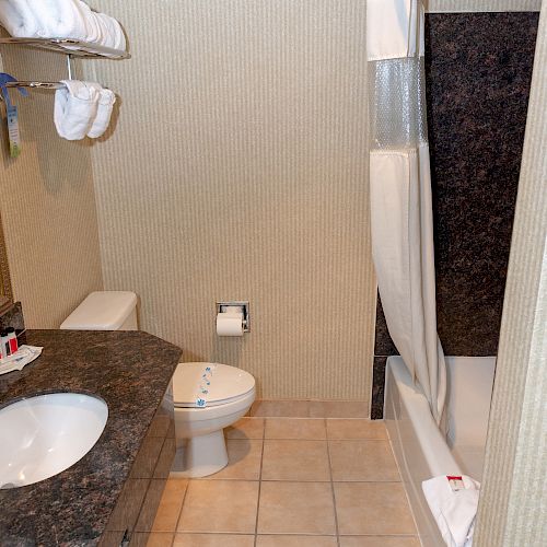 The image shows a bathroom with a sink, toilet, and a bathtub with a shower curtain. Towels are hung on a rack above the toilet and on the counter.