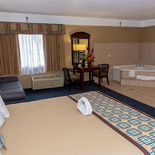 This image shows a hotel room with a large bed, sofa, table, chairs, large tub, plant, and window with curtain. The space is clean and well-lit.