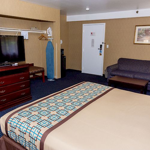 A hotel room featuring a large bed, TV, dresser, ironing board, lamp, sofa, and wall art, set on carpeted flooring with neutral-colored walls.