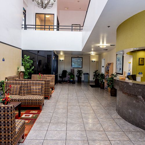 The image shows a spacious lobby area with patterned furniture, a large reception desk, indoor plants, and tiled flooring, creating a welcoming ambiance.