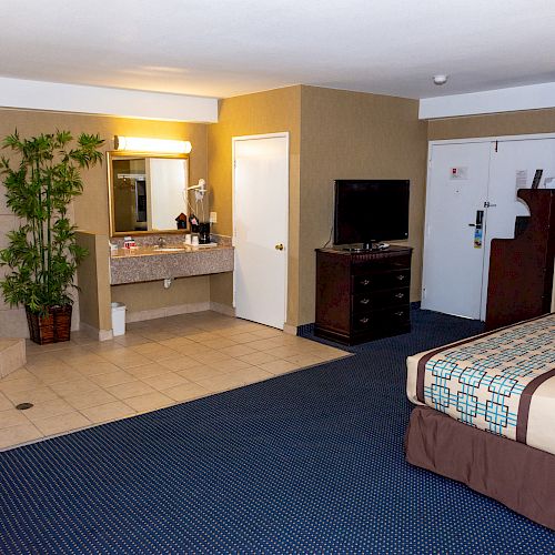 A hotel room with a bed, TV, dresser, an open bathroom area, a table, a chair, and a spa bathtub in the corner.