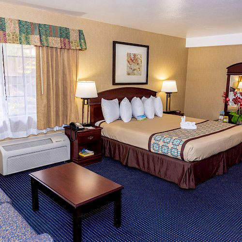 The image shows a hotel room with a bed, nightstands, lamps, sofa, coffee table, air conditioner, wall art, flowers on a desk, and a window.