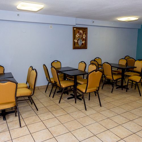 This image shows a room with several tables and chairs arranged neatly on tiled flooring, featuring a painting on the wall and overhead lighting.