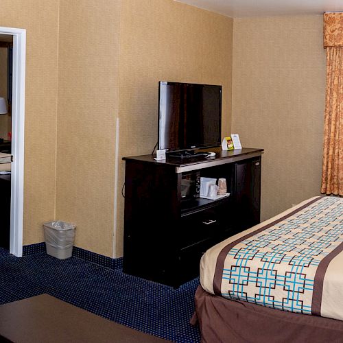 A hotel room with a bed, sofa, TV, desk, and bathroom. The room has beige walls, blue carpet, and brown and orange accents, including curtains and bedding.