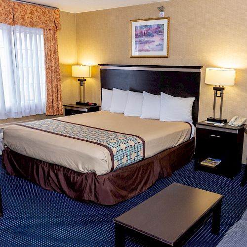 A comfortable hotel room with a large bed, nightstands, lamps, a window, a TV, and a seating area, decorated in neutral colors.