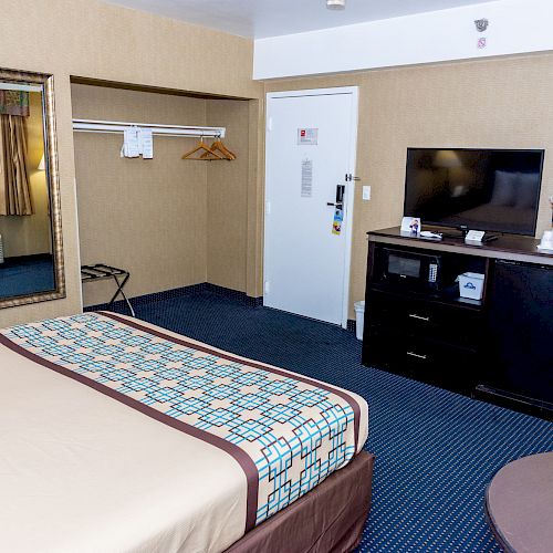 A hotel room with a bed, TV, dresser, mirror, table, and an open closet with hangers and a luggage rack, offering a basic and functional setup.