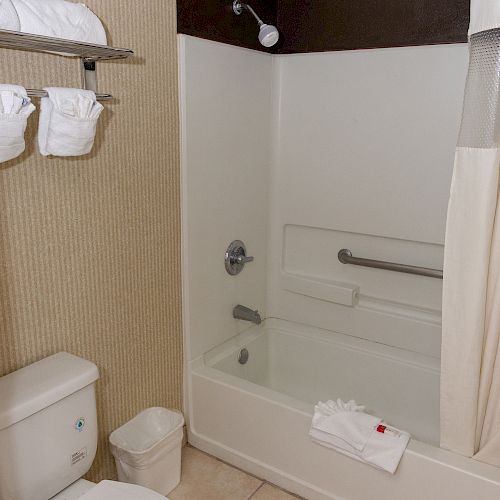 A bathroom features a toilet, a bathtub with a shower, a curtain, towel racks with towels, and a small waste bin.