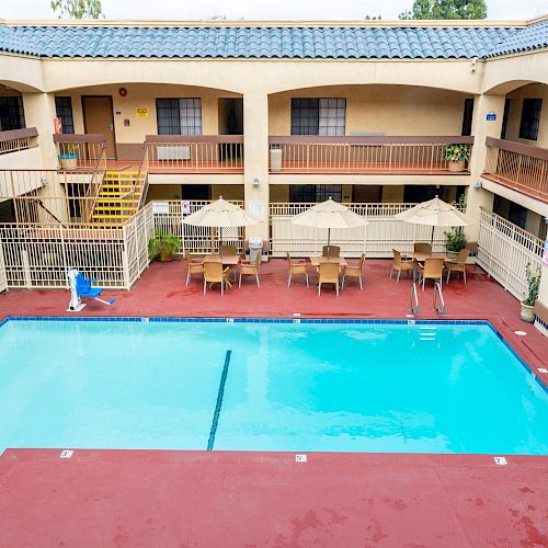 An outdoor hotel pool area surrounded by two-story beige buildings with balconies. There's a red deck, lounge chairs, and patio umbrellas.