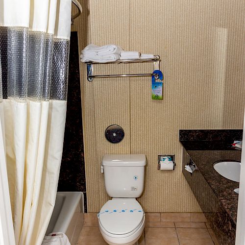 A bathroom with a shower, toilet, and sink. Towels are on a rack above the toilet, with beige walls and tiled floor.