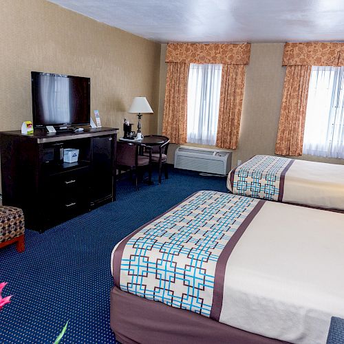 This image shows a hotel room with two beds, a TV on a dresser, a desk with a chair, an armchair, two windows, a plant, and a vibrant red flower.