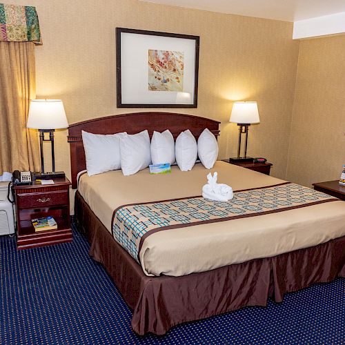 The image shows a neatly arranged hotel room with a large bed, nightstands, lamps, a dressing table with flowers, and air conditioning under the window.