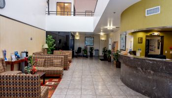 The image shows a lobby with a reception desk on the right, seating area on the left, and plants and decor throughout the space.