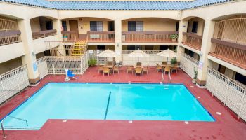 The image shows an outdoor swimming pool in a courtyard with surrounding two-story motel-style accommodations and several poolside tables and chairs.