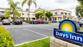 A parking lot with multiple cars in front of a Days Inn hotel, surrounded by palm trees and greenery, under a partly cloudy sky.