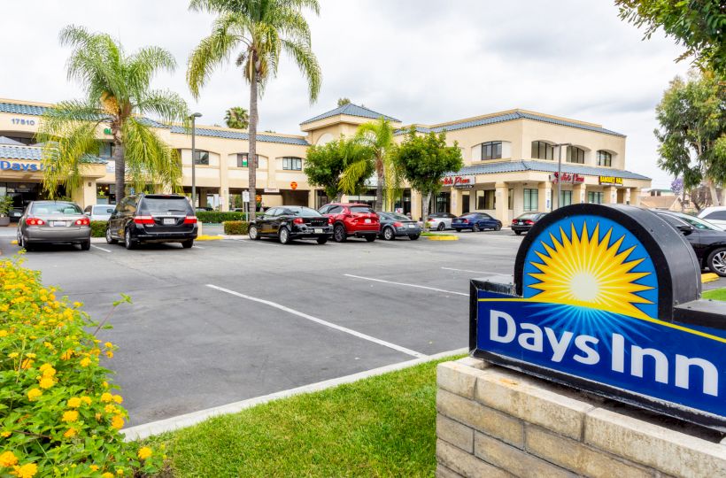 A parking lot with multiple cars in front of a Days Inn hotel, surrounded by palm trees and greenery, under a partly cloudy sky.