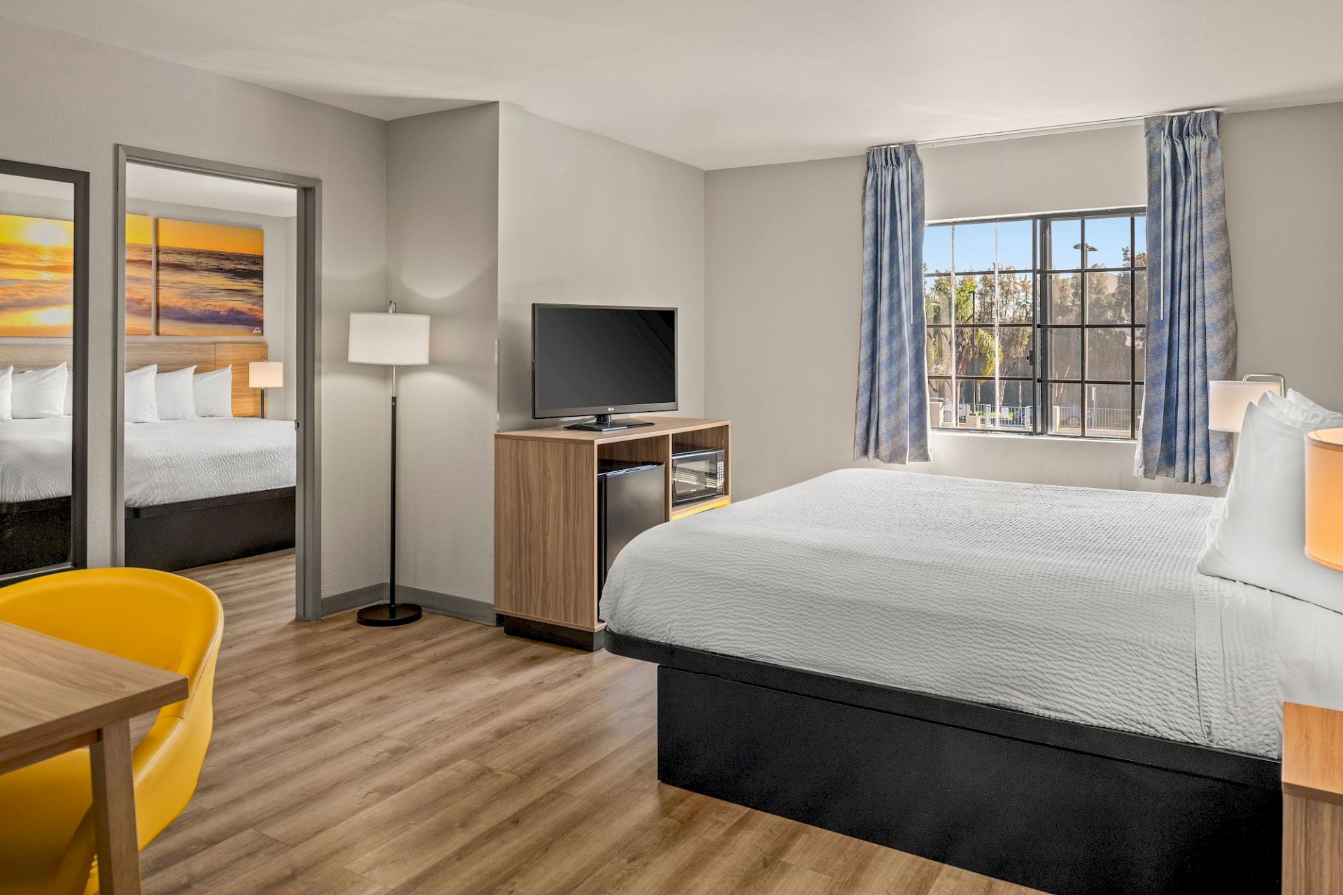 The image shows a modern hotel room with a bed, lamp, TV, and a window with curtains. There's also a second bed visible through an open door.