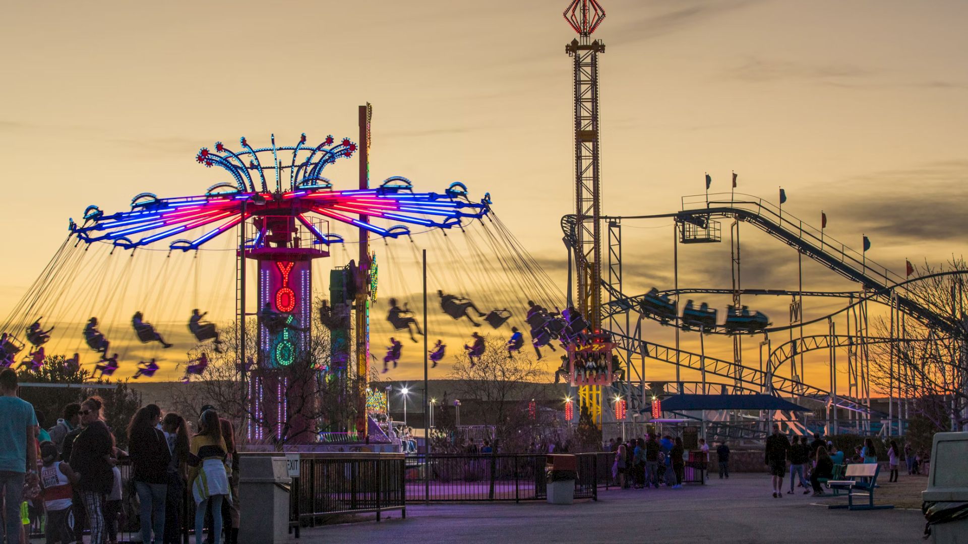 An amusement park at sunset with a swinging ride and roller coaster, lit with colorful lights, and people enjoying the attractions.