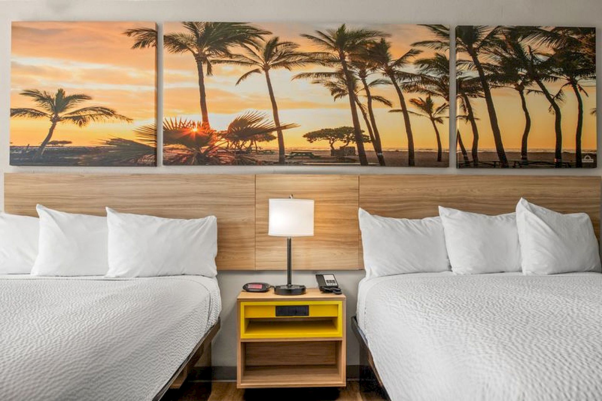 The image shows a hotel room with two beds, a central nightstand, a lamp, and a wall art of a palm tree sunset above the headboards.