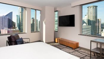 A modern hotel room with city views, featuring a bed, wall-mounted TV, a chair, and large windows.