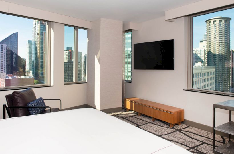 A modern hotel room with city views, featuring a bed, wall-mounted TV, a chair, and large windows.