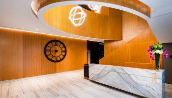 A modern interior with a marble reception desk, wooden accent walls, large clock, spiral staircase, and floral arrangement.