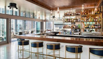 A modern bar with stylish barstools, a wooden counter, and a well-stocked shelves, with large windows offering a city view in the background.