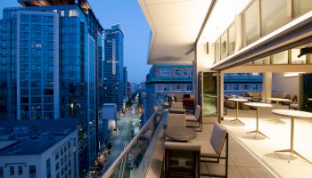 A modern cityscape with tall buildings and a well-lit balcony. The balcony features seating areas and tables, overlooking a busy street below.