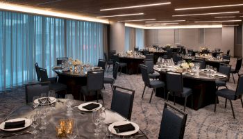 The image shows an elegant dining hall set up for an event, with round tables, black chairs, neatly arranged table settings, and dim lighting.