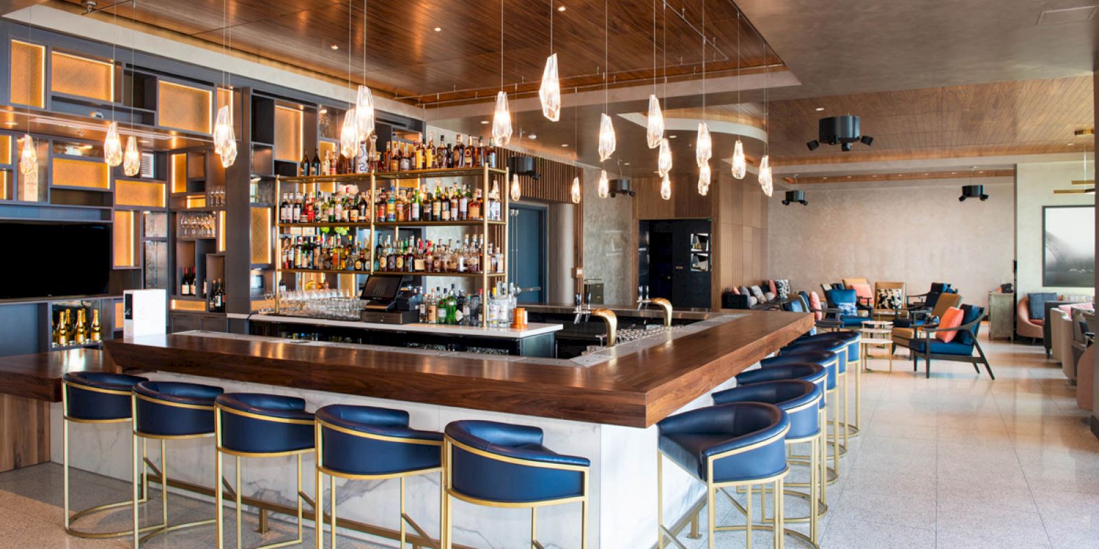The image shows a modern bar with a wooden countertop, surrounded by blue stools, featuring hanging lights and shelves stocked with various bottles of alcohol.