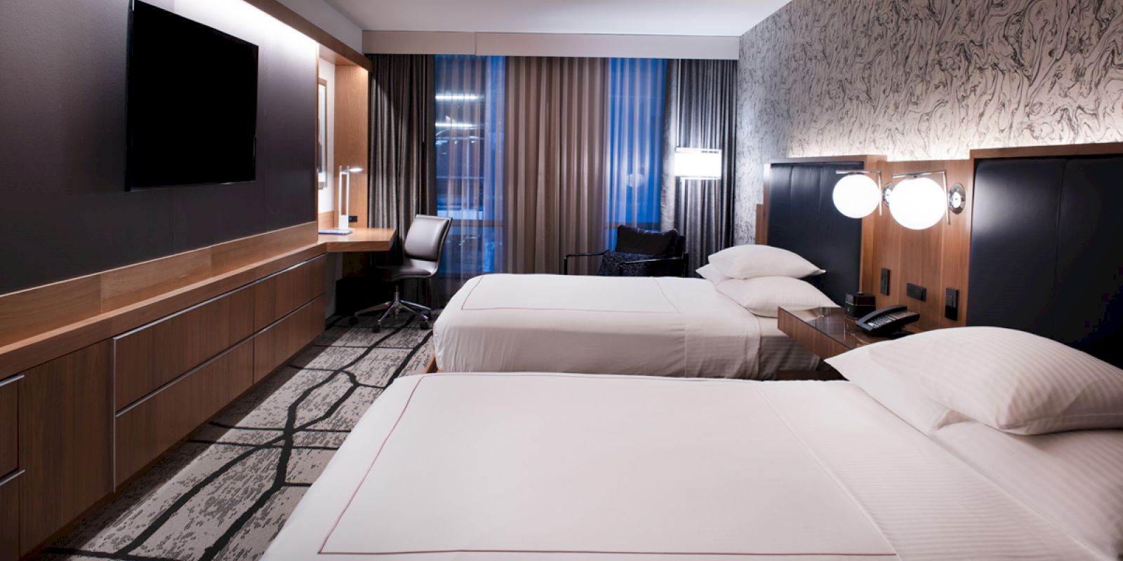 A modern hotel room with two neatly made beds, a flat-screen TV, a work desk with a chair, and stylish decor.