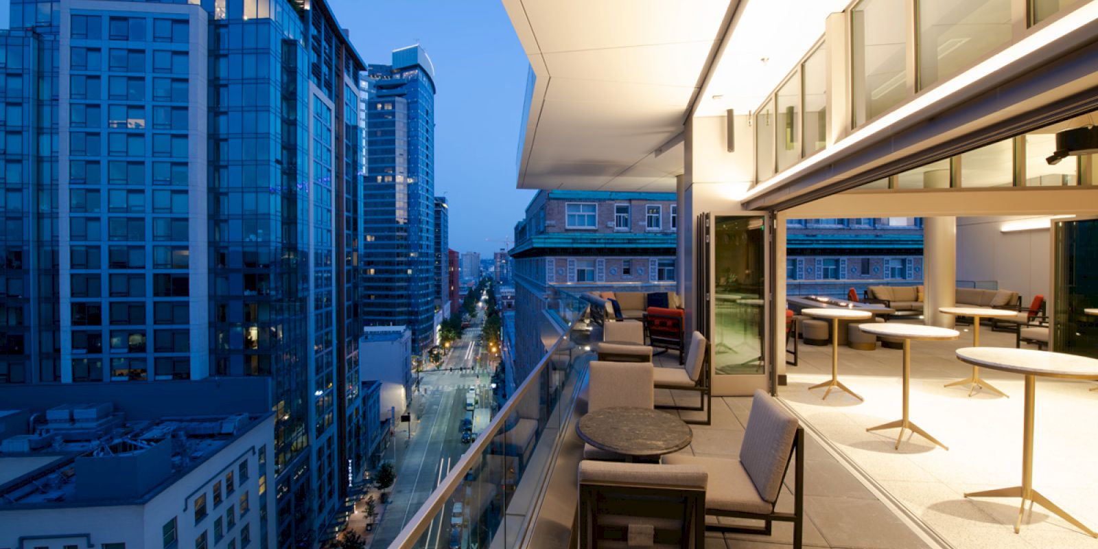A modern balcony with outdoor seating overlooks a city street flanked by tall buildings, with a spacious indoor area visible through large glass doors.