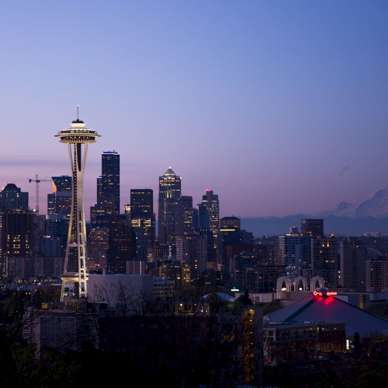 The image shows the Seattle skyline at dusk, with the iconic Space Needle prominently visible and Mount Rainier in the background.