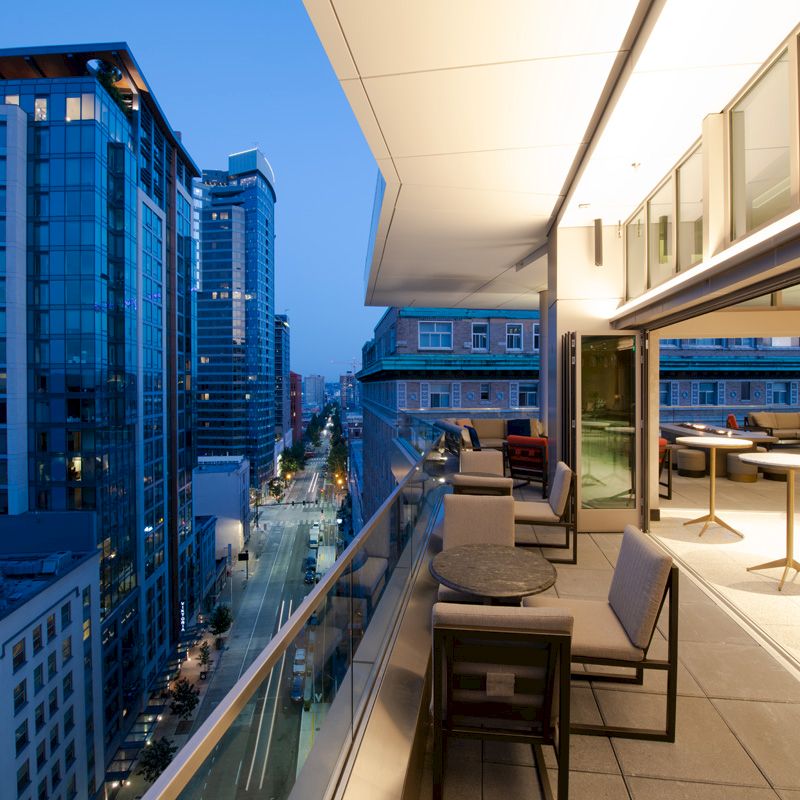 This image shows a modern cityscape at dusk, featuring a rooftop terrace with seating and tables overlooking tall buildings and a street below.