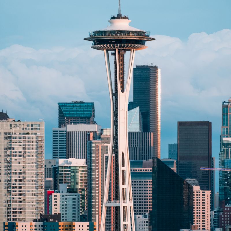 The image shows the Space Needle surrounded by buildings in Seattle's downtown area, with a partly cloudy sky in the background.