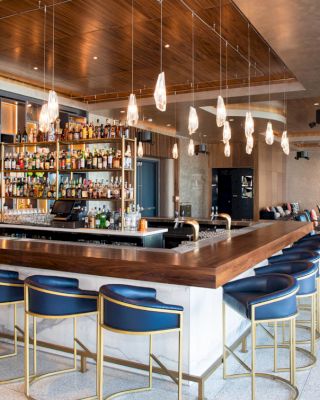 The image shows a modern bar with a wooden counter, blue bar stools, a variety of drinks on shelves, and stylish pendant lights, ending the sentence.