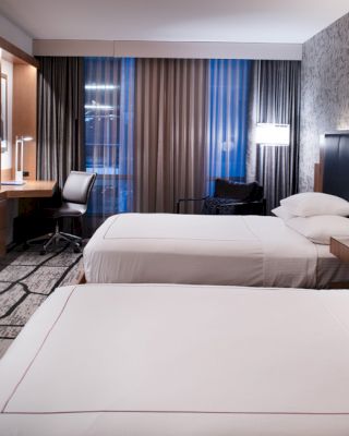 A modern hotel room with two beds, a TV, a desk, and a window with blinds and curtains. The walls have a decorative design and lighting.