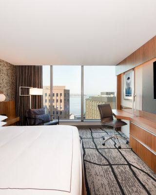 This image shows a modern hotel room with a large bed, a desk with a chair, a flat-screen TV, and a window with city and water views.