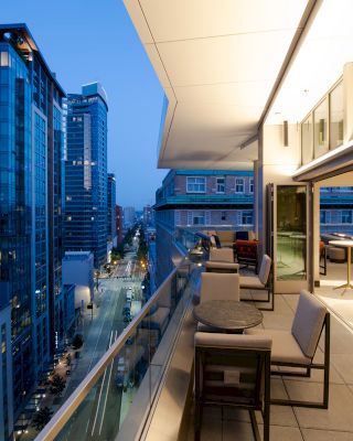 The image depicts a modern high-rise apartment balcony view of a cityscape at dusk, with outdoor seating, glass railings, and adjacent buildings.
