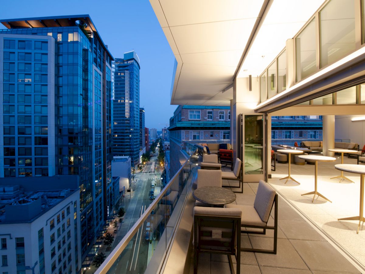A stylish rooftop terrace with seating overlooks a cityscape of tall modern buildings during early evening, showcasing a vibrant urban environment.
