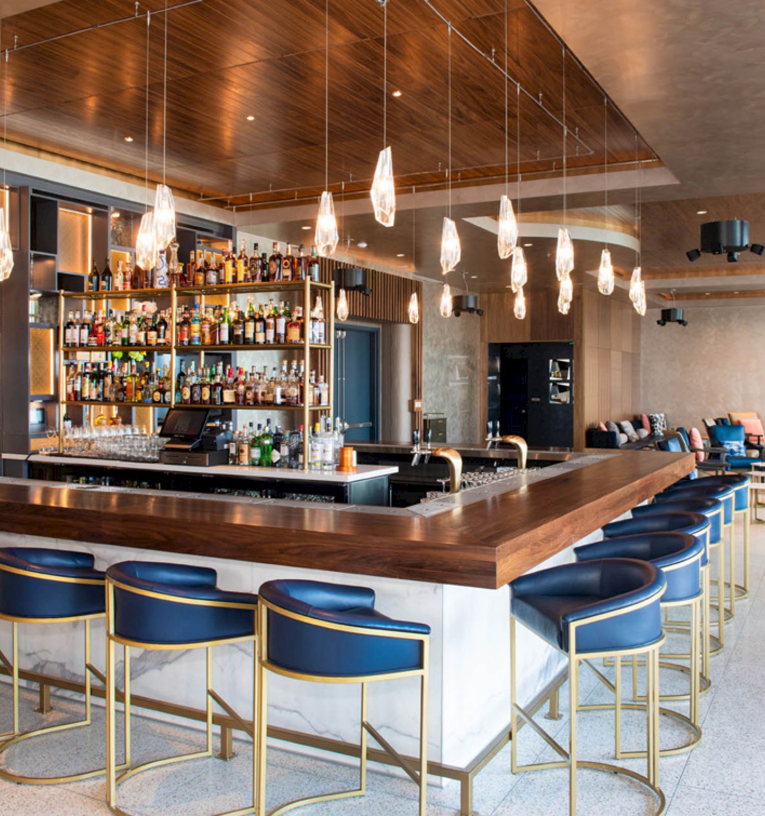 This image shows a modern bar with a wooden countertop, blue chairs, various bottles on shelves, and hanging pendant lights in a stylish interior.
