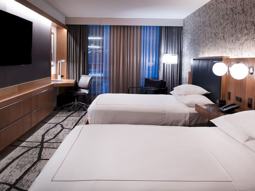 A modern hotel room with two beds, a wall-mounted TV, desk, and chair near the window with curtains, and two lamps on the nightstand ending the sentence.